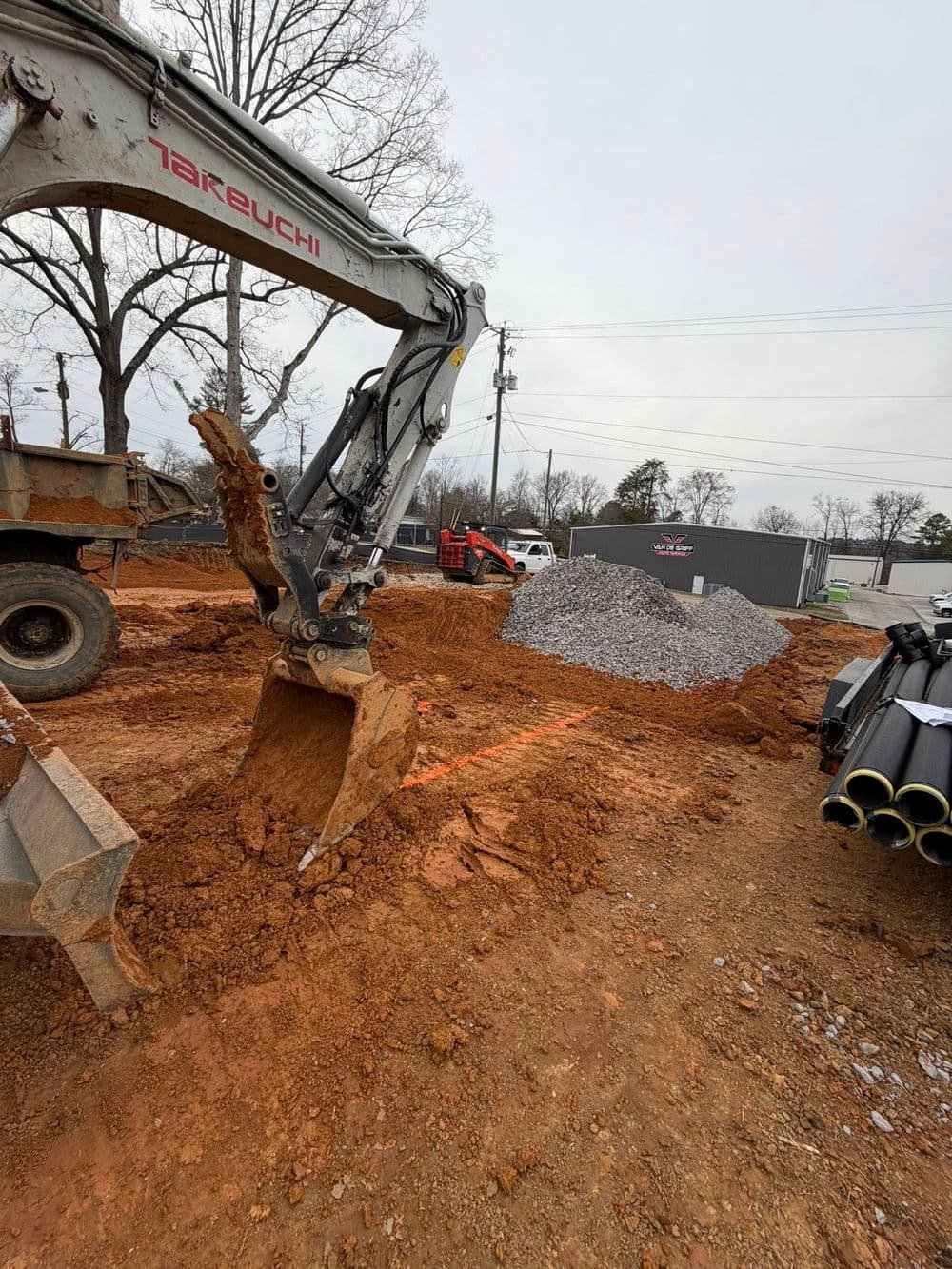 Excavator digging in ground, piles of gravel and construction materials on site.