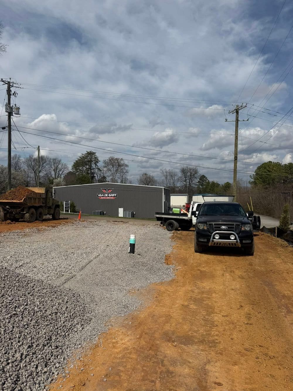 Construction site with gravel road, trucks, and a gray building in the background under cloudy sky.