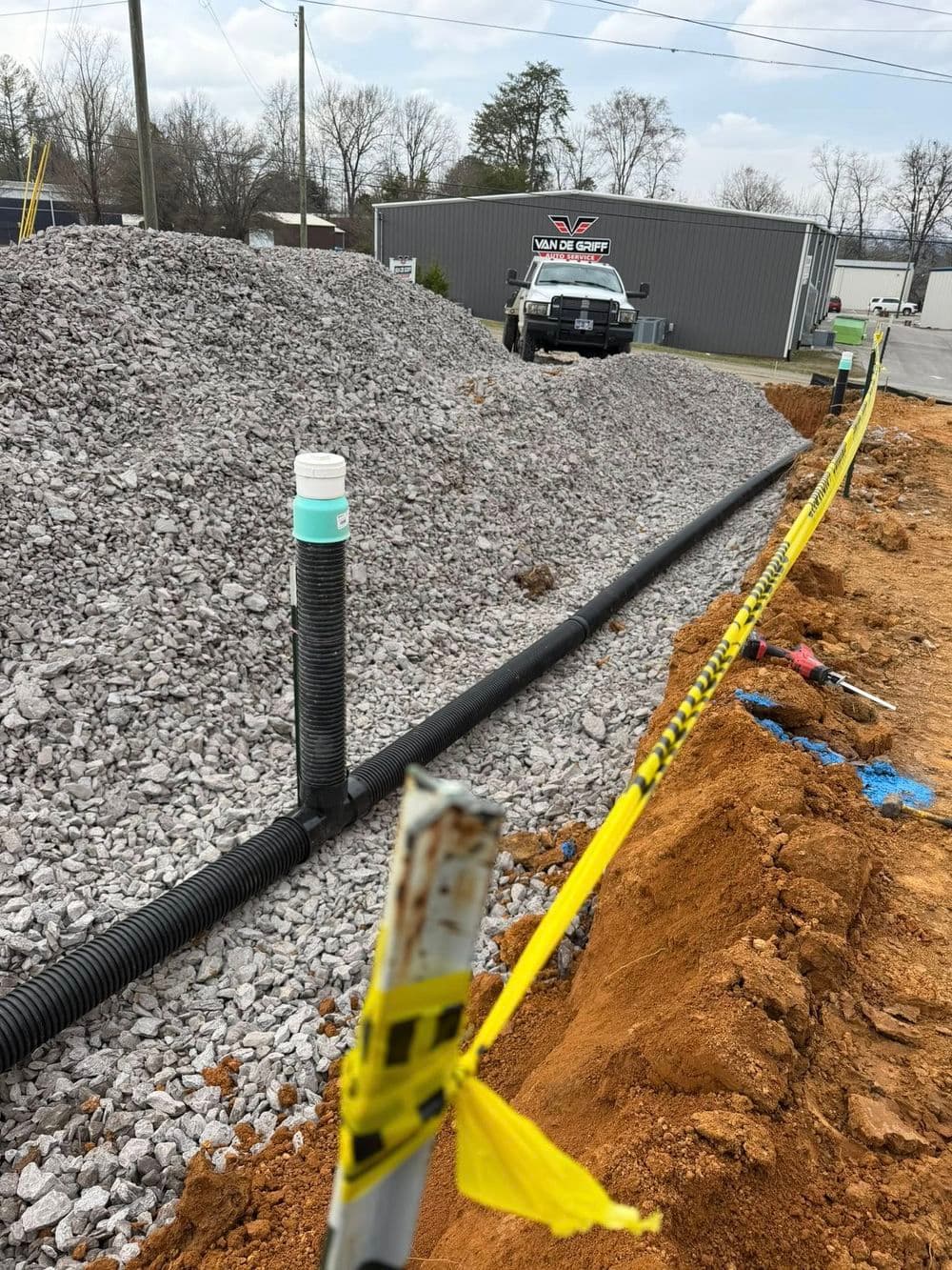 Construction site showing gravel pile, drainage pipe, and yellow caution tape.