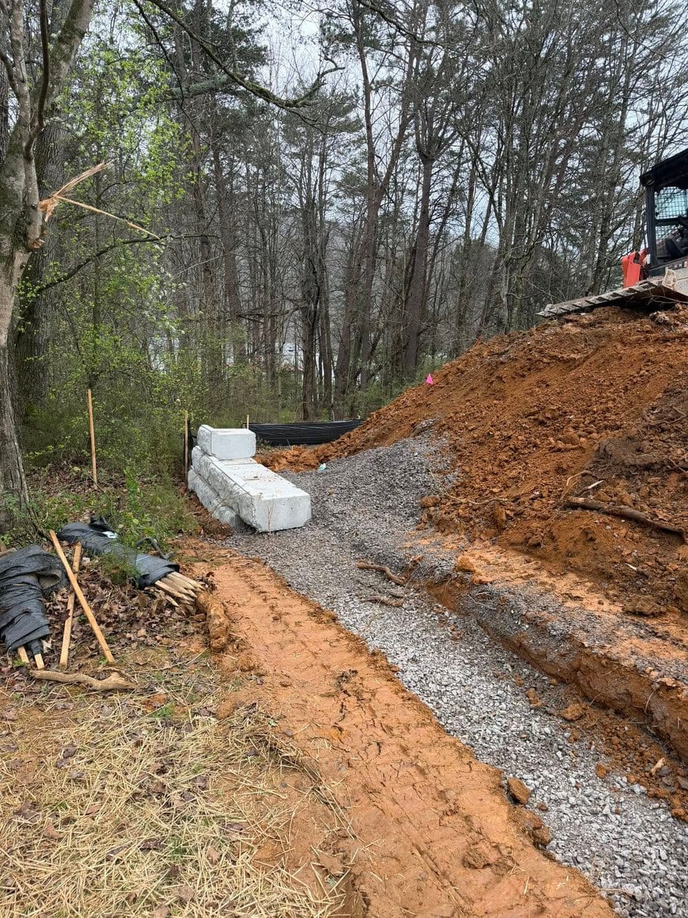 Construction site with a gravel path, earthwork, and retaining wall among wooded area.