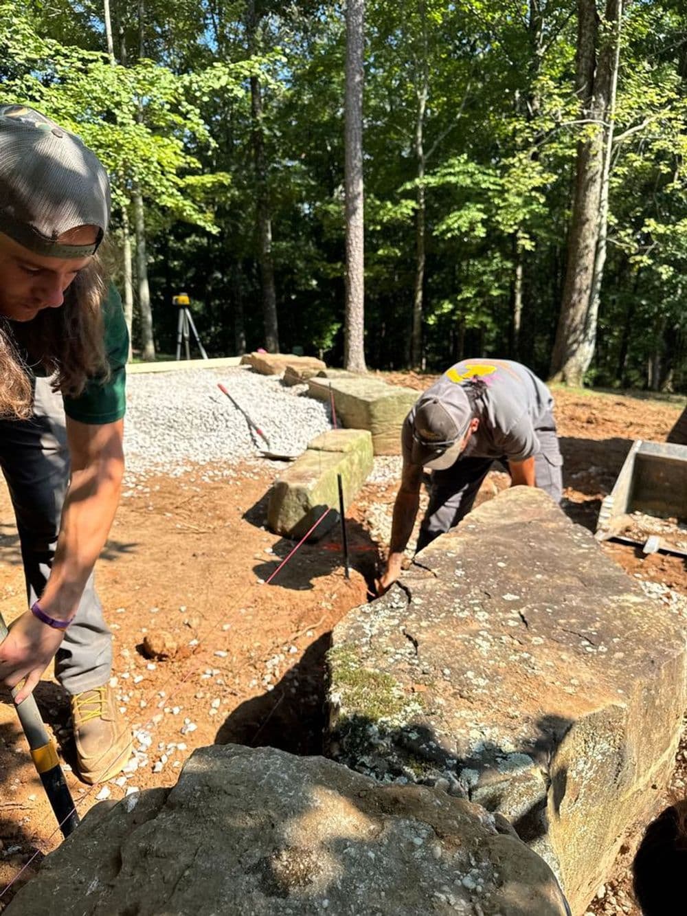 Two workers excavating rocks at a construction site surrounded by trees.