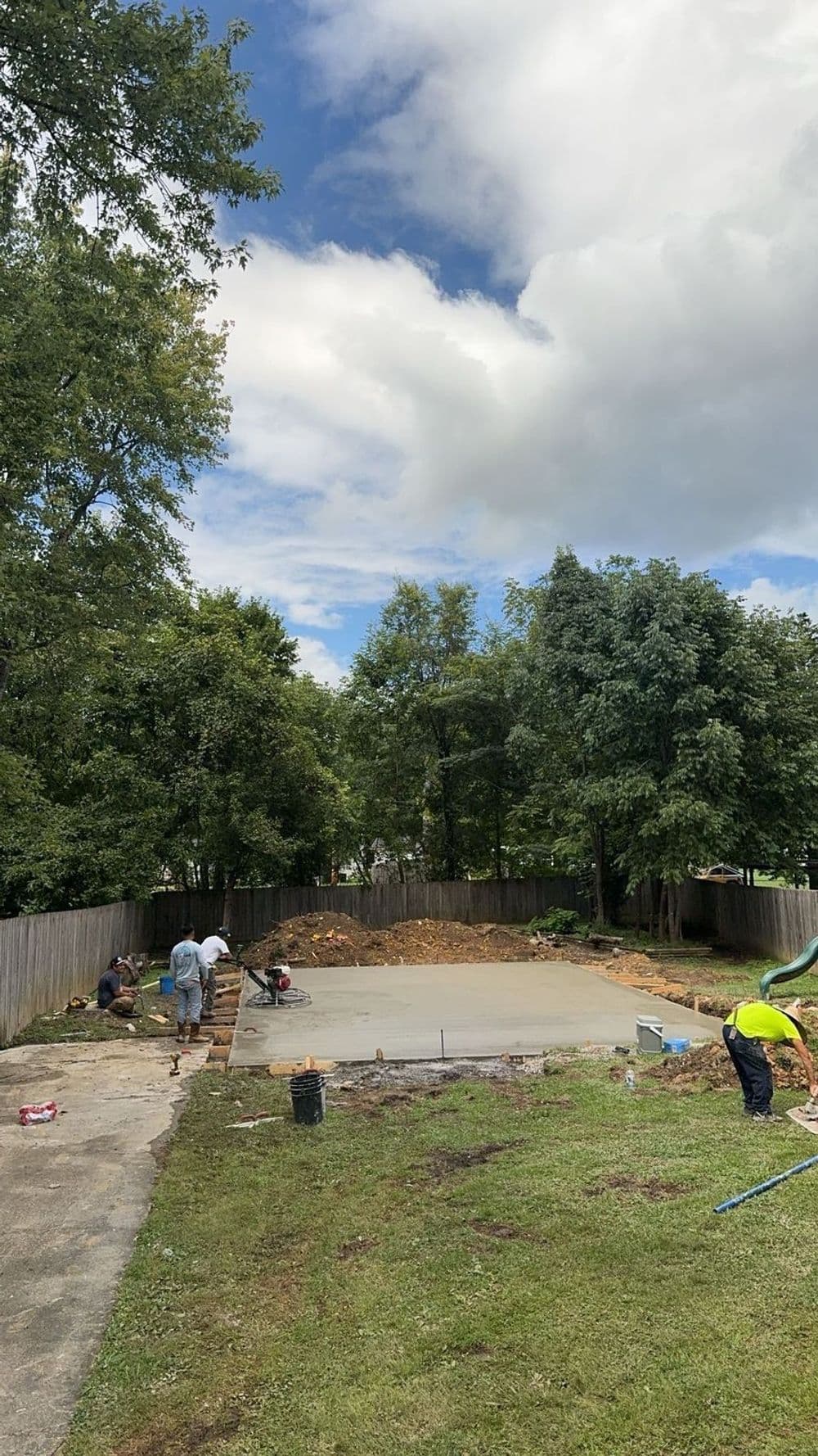 Construction workers preparing a concrete slab in a residential backyard under a cloudy sky.