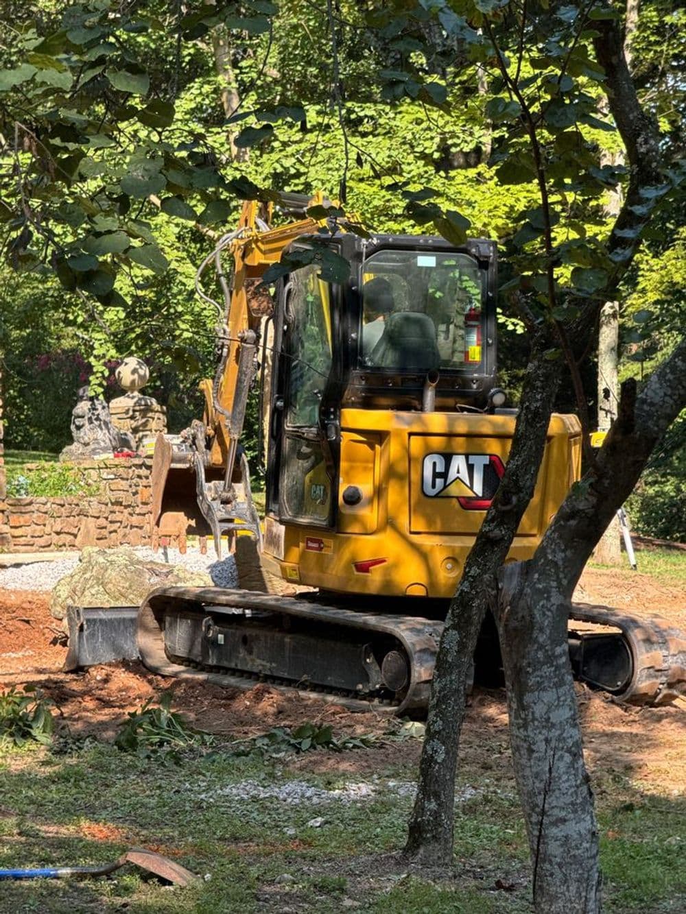 Yellow CAT excavator working in a landscaped area surrounded by trees and greenery.